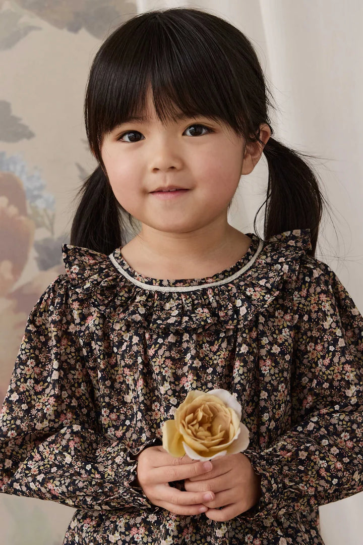 A young girl with dark hair in pigtails wearing a long-sleeve black floral dress, holding a beige rose.