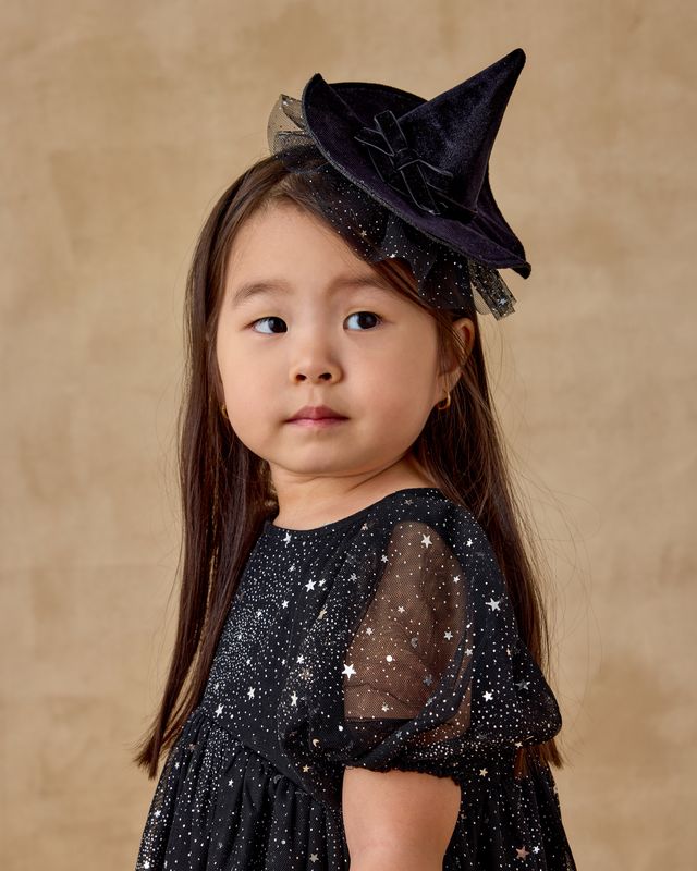 Young girl wearing a black witch hat and dress with star patterns on a beige background