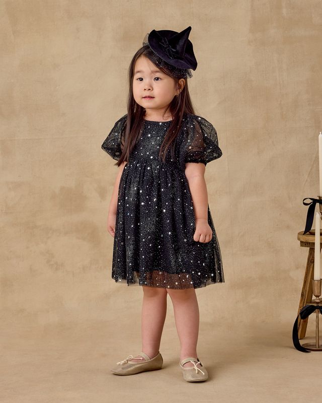 Young girl wearing a black star-patterned dress and hat against a beige background