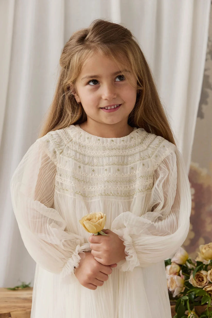 A young girl with long light brown hair wearing a white smocked blouse dress with sheer long sleeves, holding a small yellow rose against a soft white curtain background.
