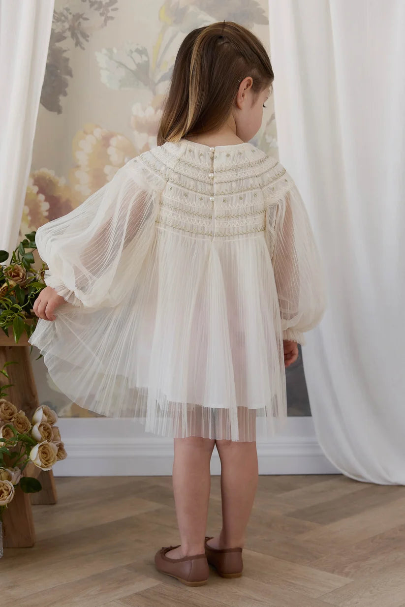 A young girl viewed from behind wearing a cream-colored tulle dress with embroidered detailing on the back, standing in a room with a floral wallpaper background and white curtains.