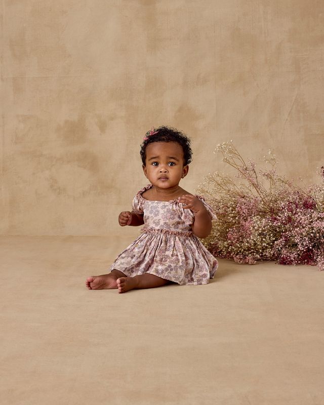 Young child in a floral dress sitting on a beige floor with dried flowers.