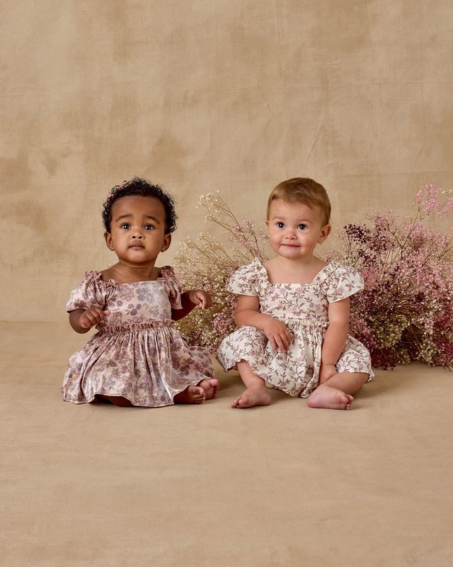 Two young children in floral dresses sitting on a beige surface with flowers in the background.