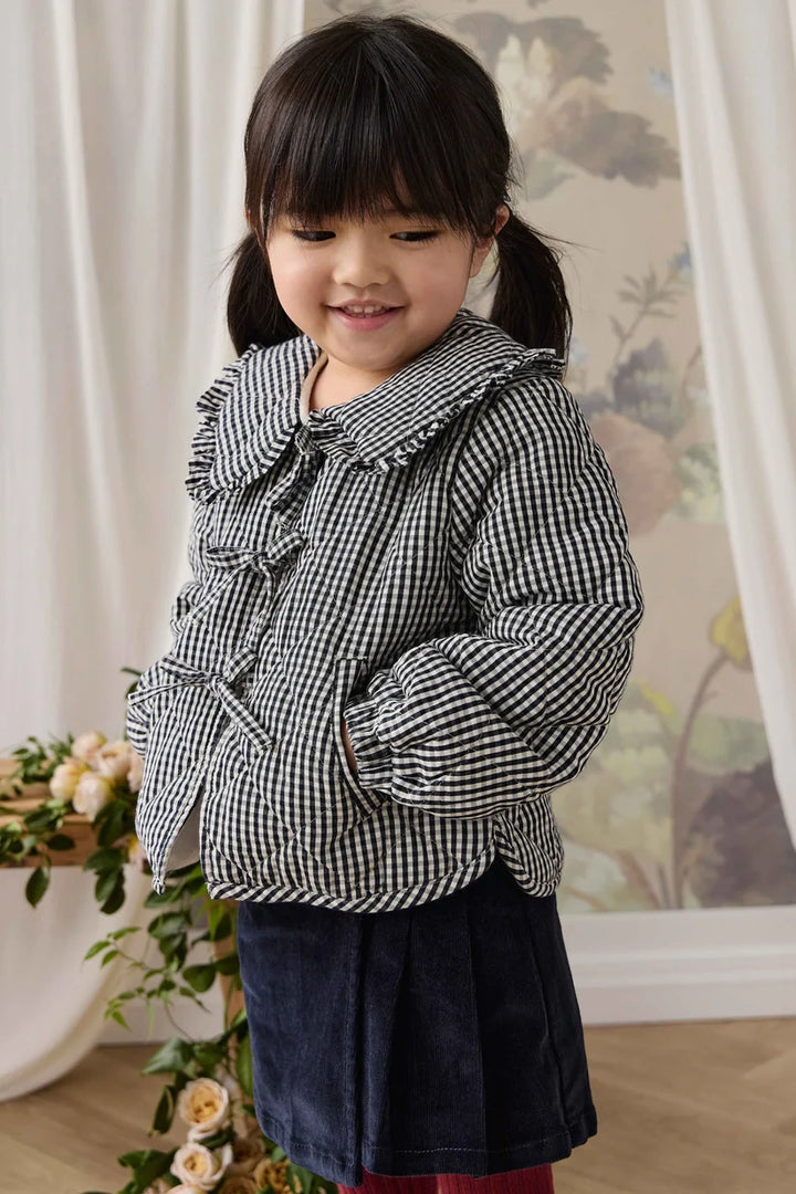 A young girl with dark hair in pigtails wearing a black and white gingham long-sleeve jacket with ruffles and a dark navy skirt, standing in a styled indoor setting with curtains and a floral print background.