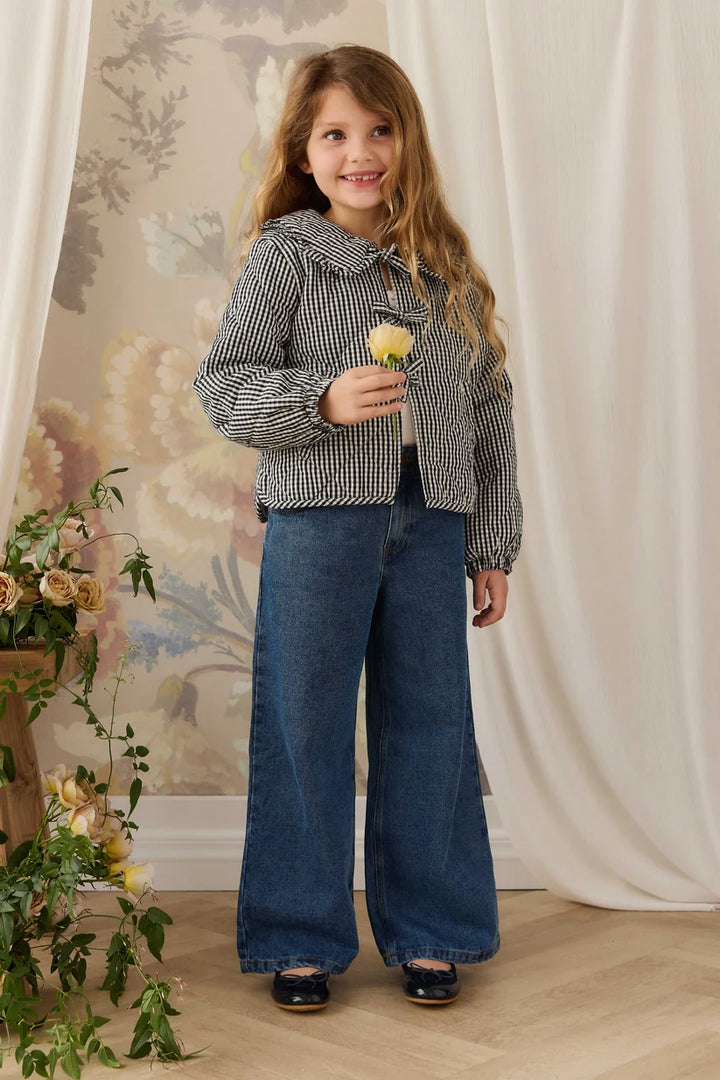 A young girl stands indoors wearing a black-and-white patterned cropped jacket, wide-leg blue jeans, and dark shoes, holding a small yellow flower. The room has cream draped fabric, a floral wall mural, and a light wood floor.