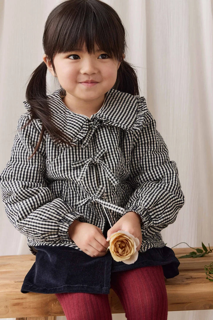 A young girl with dark hair in pigtails sits on a wooden bench wearing a black and white gingham long-sleeve jacket with a ruffled collar and bow, a dark skirt, and red tights, while holding a beige rose.