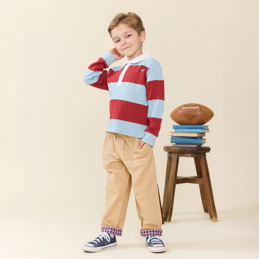 A young boy stands in a studio wearing a long-sleeve rugby-style shirt with red and light blue horizontal stripes, tan pants, and navy sneakers. A small wooden stool holds a football and stacked books behind him against a plain warm beige background.