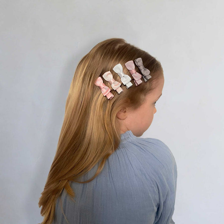 A side-profile view of a young girl with long blonde hair wearing a set of six pastel bow hair clips in pink, white, and light blue, set against a plain light gray wall.