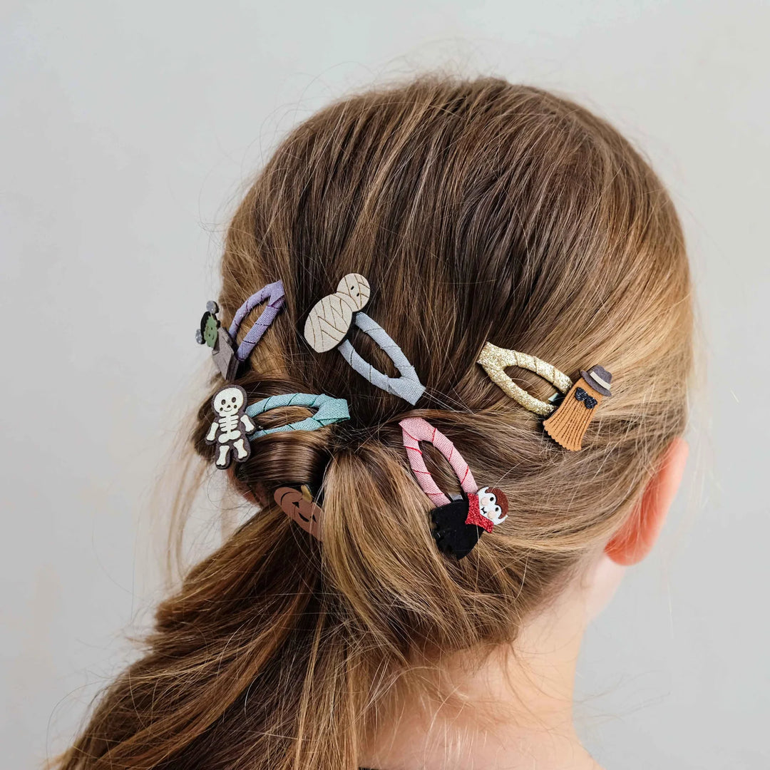 A close-up back view of a person with light brown hair styled in a low ponytail, secured with multiple colorful novelty hair clips featuring cartoon and Halloween-themed designs.