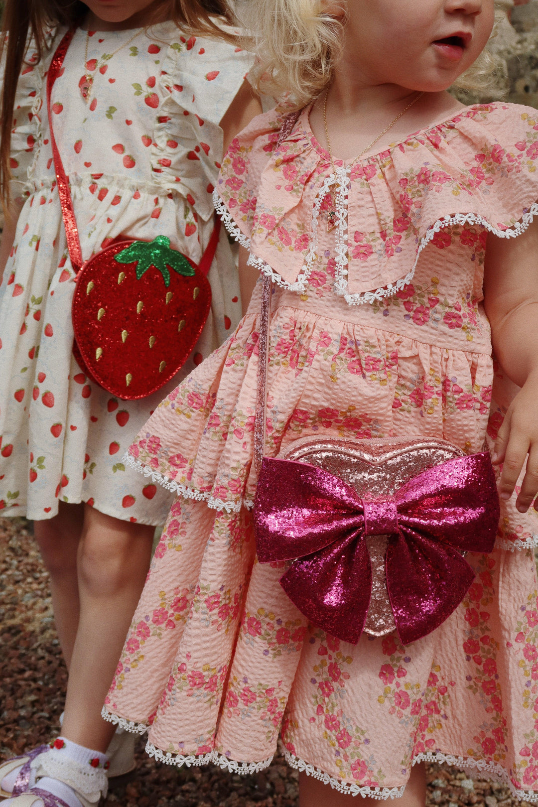 little girl modeling the pink dress with pink flowers standing next to a young girl wearing a dress with tiny strawberries on it and glitter strawberry bag. 
