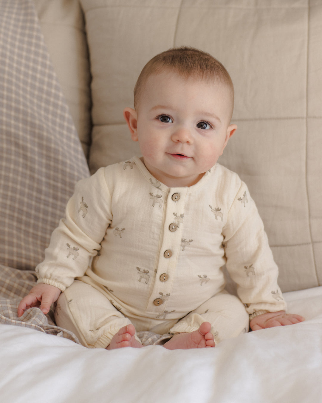 A baby sitting on a white blanket in front of a beige quilted cushion, wearing a cream-colored long-sleeve button-front romper with small moose prints.