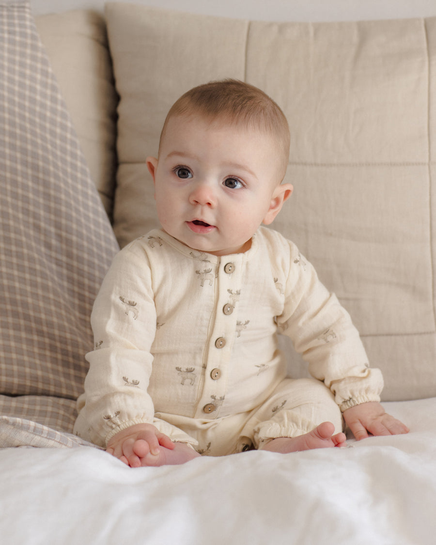 A baby sitting on a white bed wearing a cream-colored long-sleeve button-up romper with small animal prints, set against a background of beige pillows.