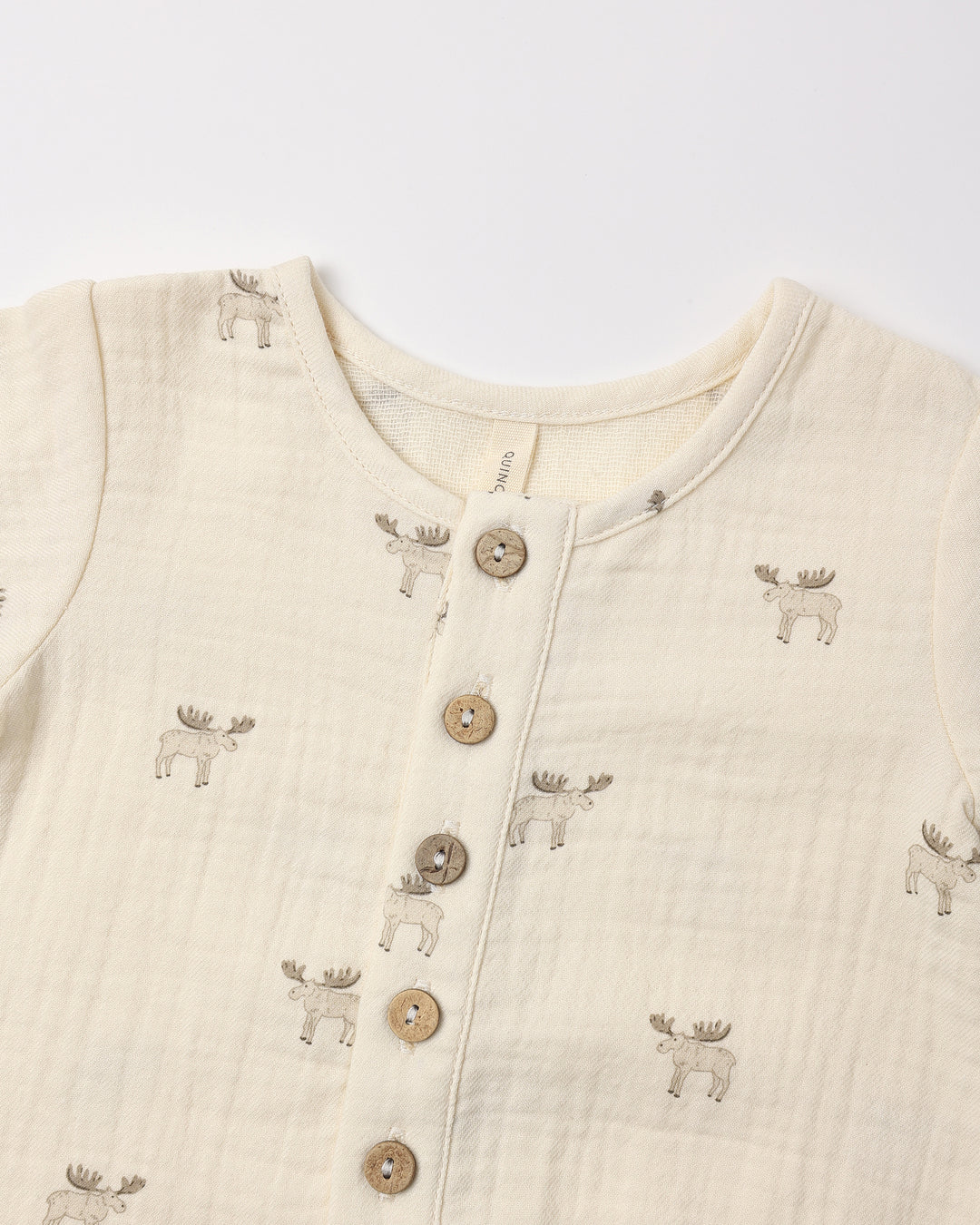 A close-up of a baby's long-sleeve muslin romper in cream, featuring a button-down front with wooden buttons and an all-over print of embroidered moose figures.