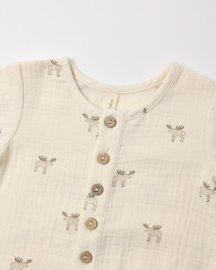 A close-up of a baby's long-sleeve muslin romper in cream, featuring a button-down front with wooden buttons and an all-over print of embroidered moose figures.