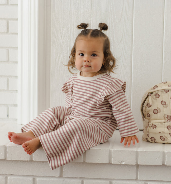 A toddler girl with two buns sits on a white brick ledge wearing a matching long-sleeve striped set. The set has thin mulberry and cream horizontal stripes.