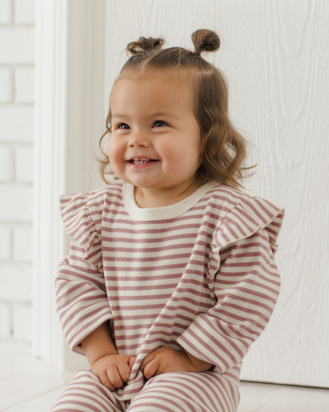 A smiling toddler with two buns sits wearing a long-sleeve ruffled shirt and matching striped pants in a soft burgundy and white stripe pattern, set against a bright white interior background.