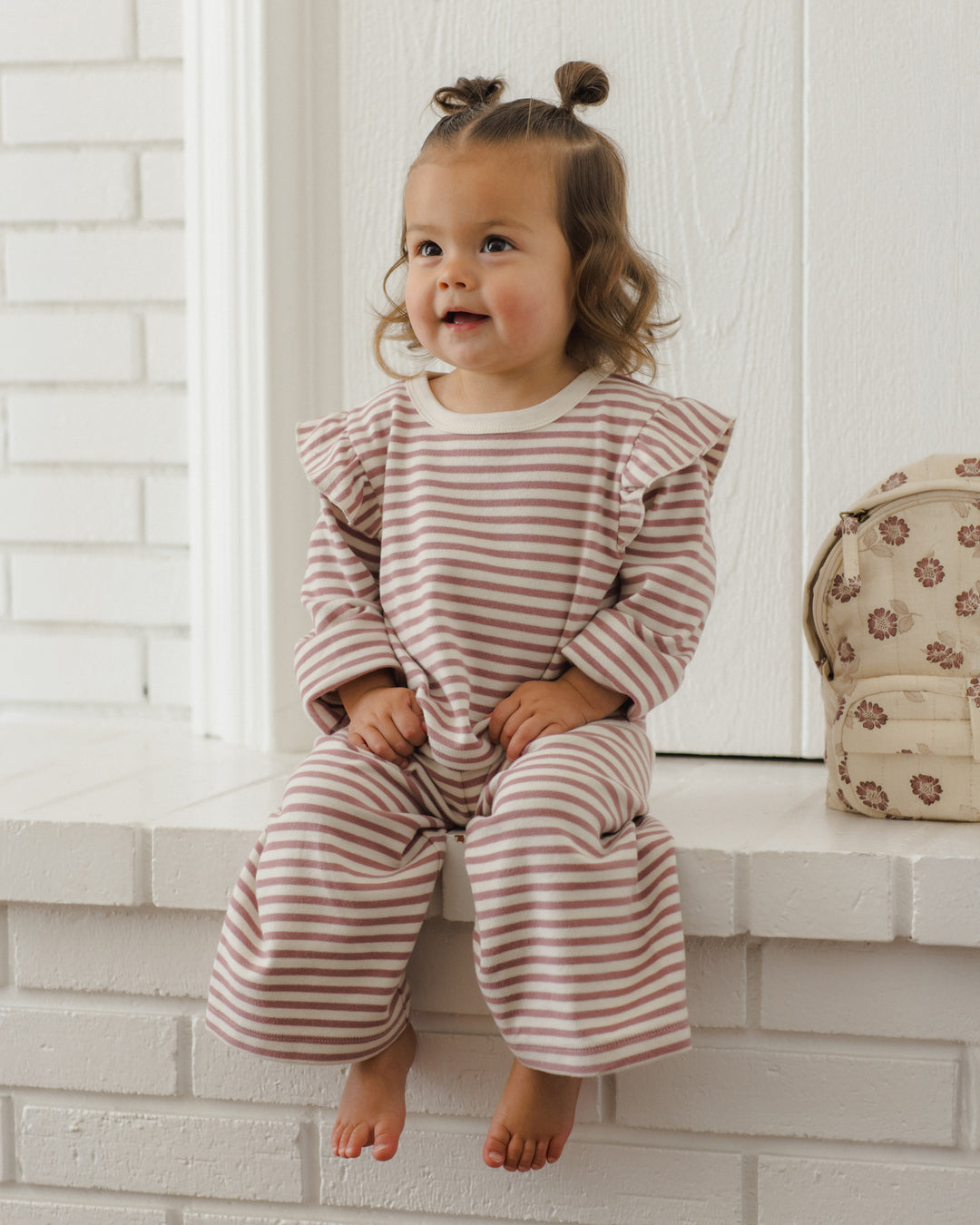 A toddler girl with two small buns sits barefoot on a white brick ledge, wearing a long-sleeve ruffled top and matching wide-leg pants in a dusty pink and white stripe pattern. A small patterned cream backpack rests on the ledge to her right.
