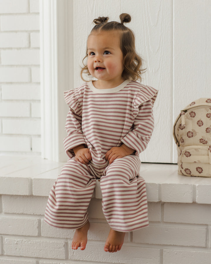A toddler girl with two small buns sits barefoot on a white brick ledge, wearing a long-sleeve ruffled top and matching wide-leg pants in a dusty pink and white stripe pattern. A small patterned cream backpack rests on the ledge to her right.