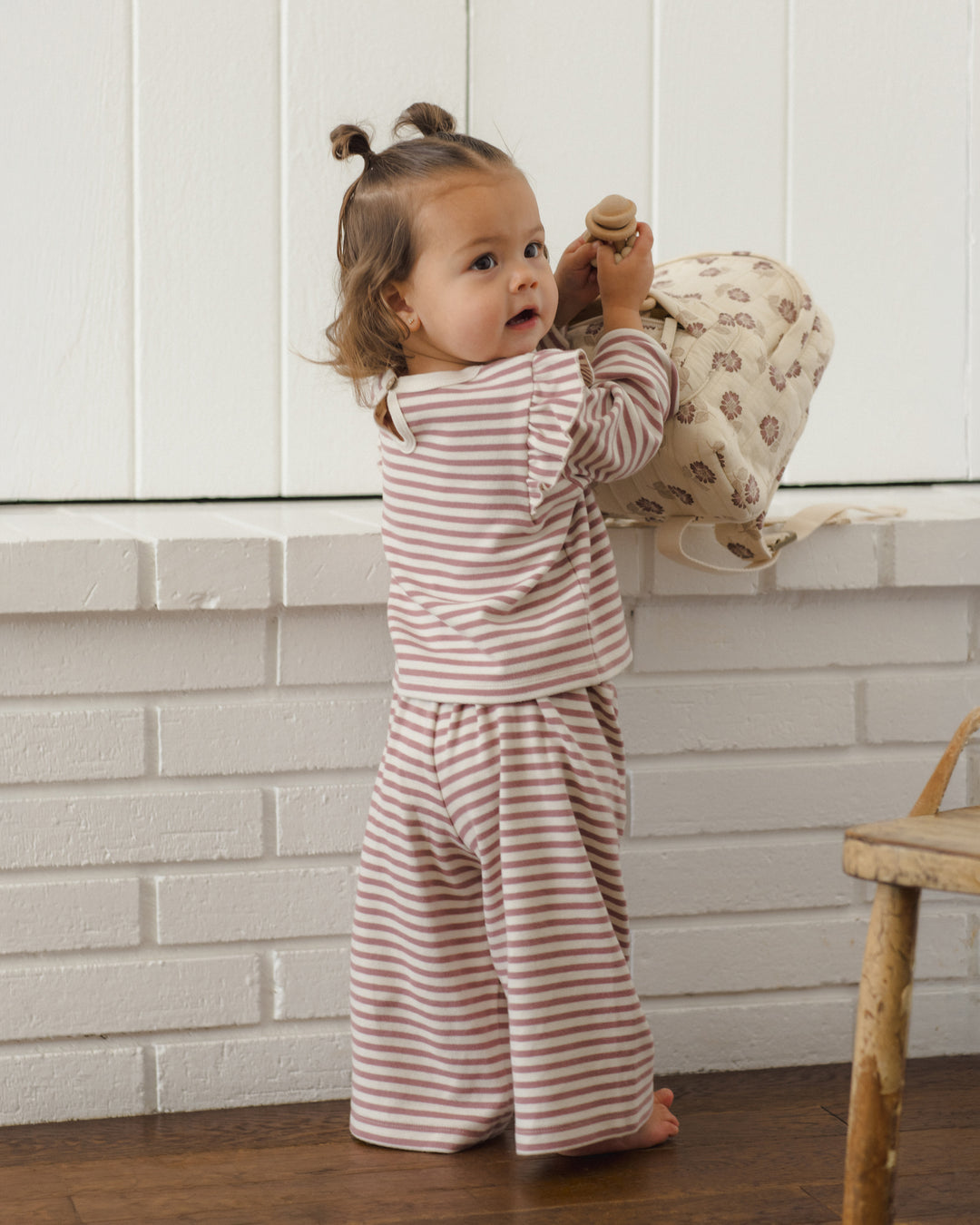 A toddler stands barefoot indoors against a white paneled wall, wearing a matching two-piece set with thin burgundy stripes on a warm off-white background. The set includes a long-sleeve top and wide-leg pants.