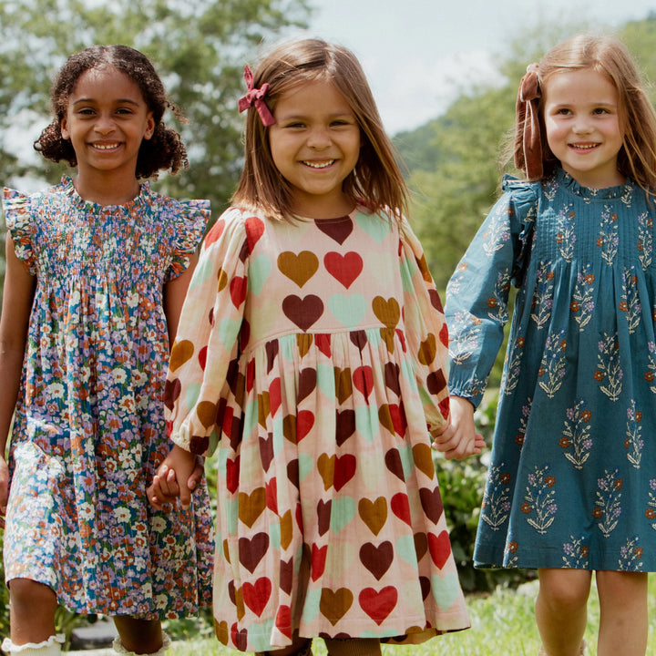 Three young girls walking hand-in-hand outdoors in a grassy field with trees and hills in the background. They are wearing long-sleeve dresses in different patterns: one blue floral dress, one cream dress with red and brown hearts, and one teal dress with a blue floral pattern.
