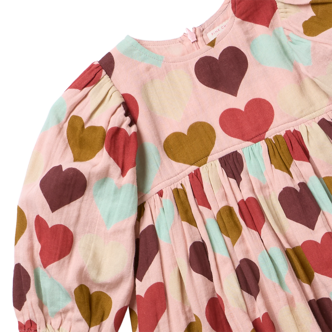 A close-up of a children's long-sleeve dress with a pink base and an allover heart print in red, mustard, and mint green. The dress features gathered detailing at the waist and sleeves, with a visible 'Miki & Co' brand label inside the neckline.