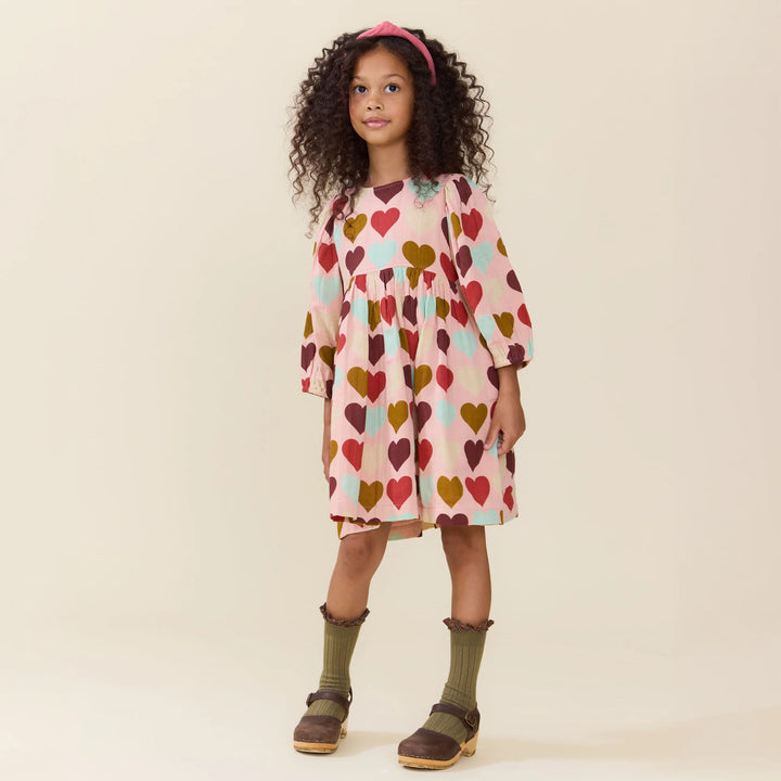 A young girl with curly hair wearing a pink long-sleeve dress with a colorful heart pattern, olive green socks, and brown clogs, standing against a solid beige background.