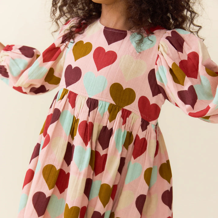 A close-up of a child wearing a pink dress with a heart pattern in red, burgundy, mint green, and mustard yellow. The dress has long puff sleeves and a gathered waist, shown against a plain cream background.