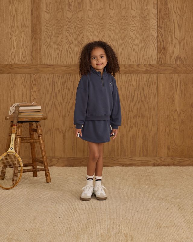 Young girl in a navy tracksuit standing in front of a wooden paneled wall.