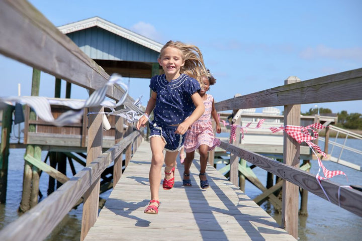 Two young girls running on a wooden dock by the water with a clear blue sky.