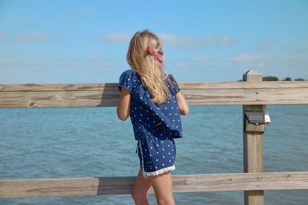 young girl in a blue polka dot dress standing on a wooden pier by the water.