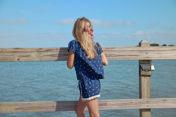 young girl in a blue polka dot dress standing on a wooden pier by the water.