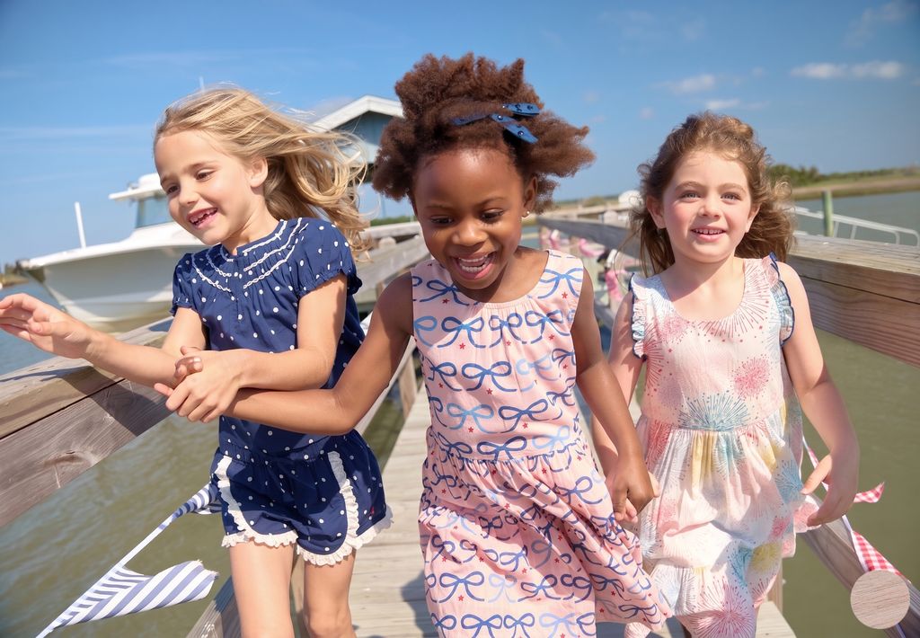 Three young girls running on a dock by a body of water with a boat in the background.