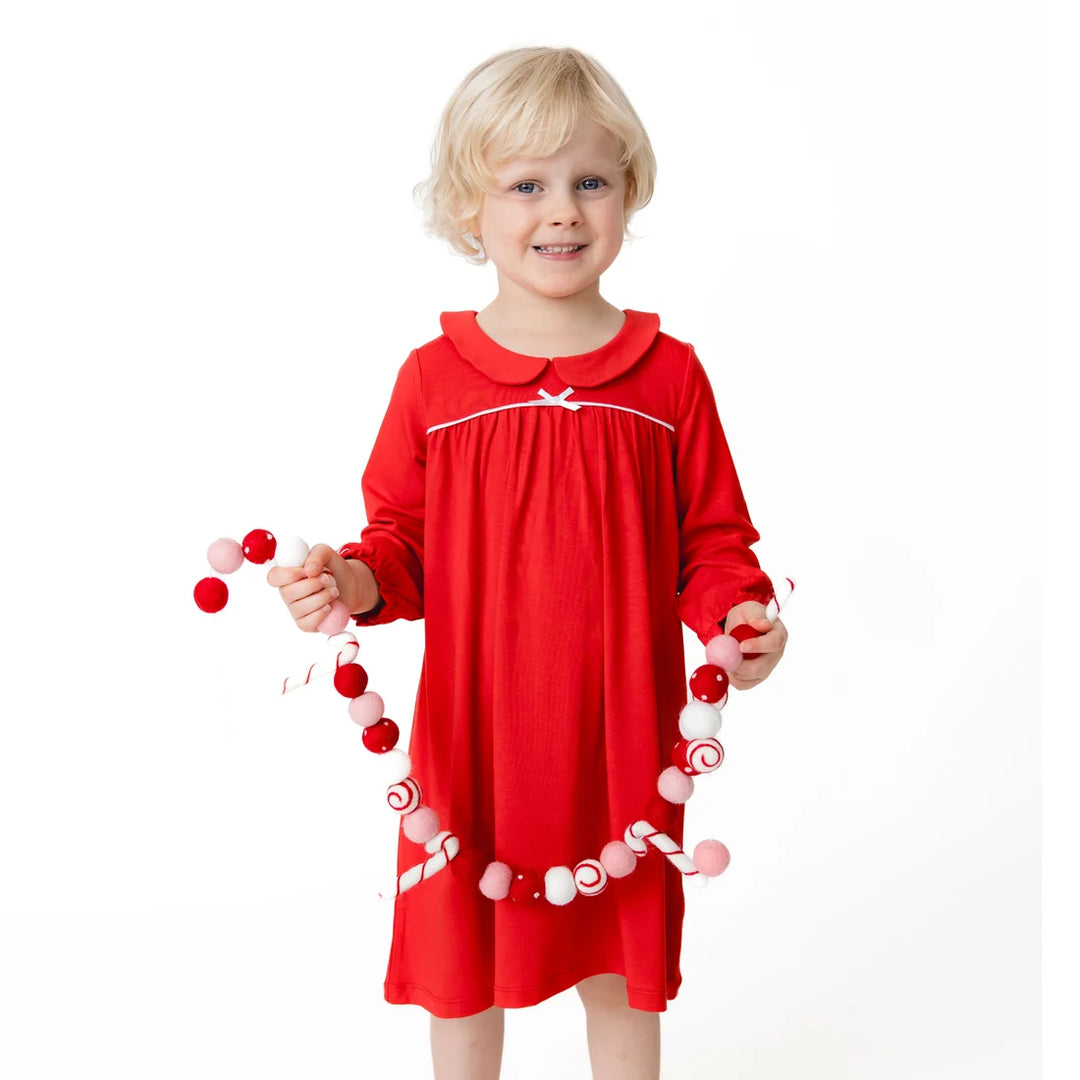 A young blonde child wearing a red long-sleeve dress with white trim, holding a red, white, and pink pom-pom garland against a plain white background.