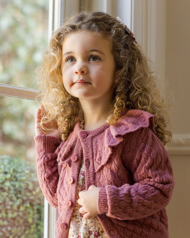 Young girl wearing a  knitted burgundy cardigan standing by a window.