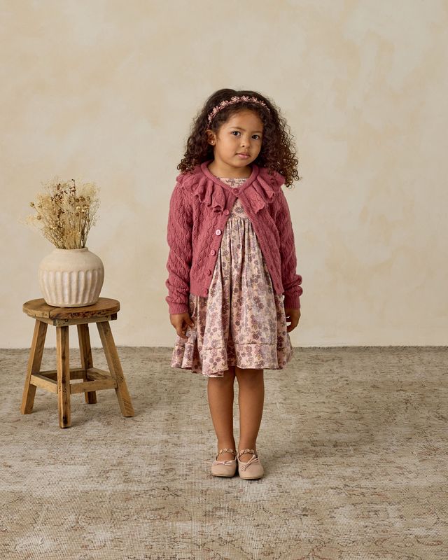 Young girl in a pink dress and pointelle cardigan standing next to a wooden stool with a vase on it.