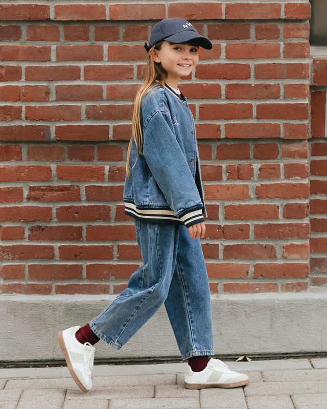 Child wearing a denim outfit with a cap, standing against a brick wall.