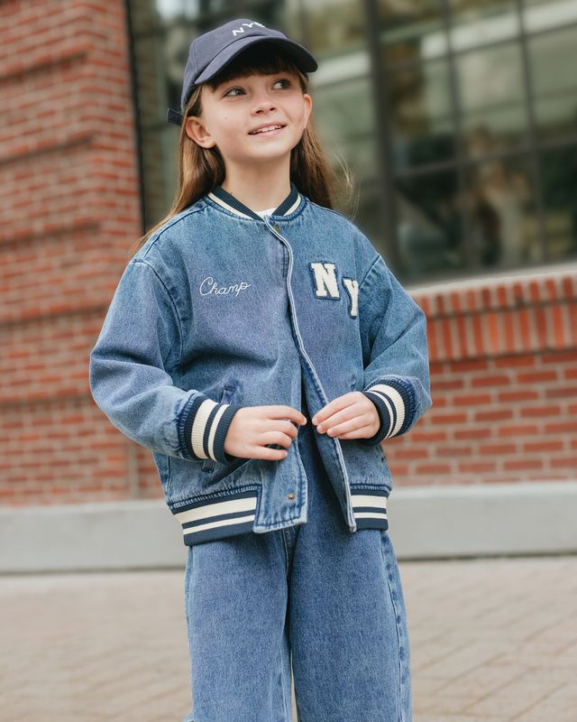 Child wearing a blue denim jacket with 'Champ' and 'NY' embroidery, standing outdoors.