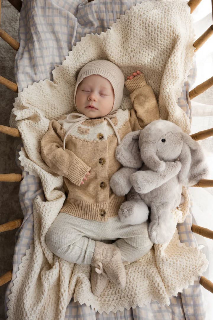 Baby in a crib with a plush elephant toy, wearing a beige outfit.