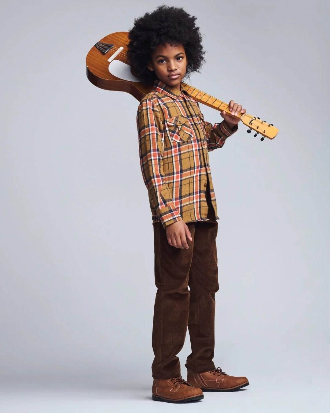 A child model with an afro hairstyle stands in profile holding a wooden acoustic guitar over their shoulder. They are wearing a brown and orange plaid flannel shirt, brown corduroy pants, and brown suede boots, set against a solid light gray studio background.