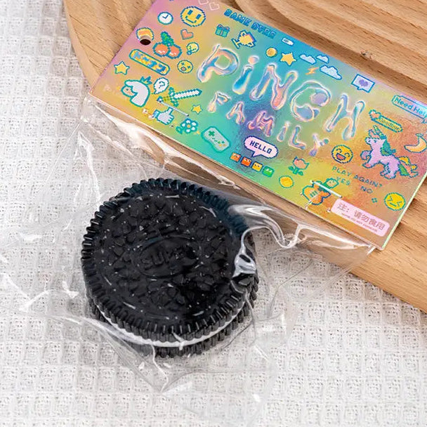 Black textured round cookie that looks like oreo in clear packaging with colorful 'Pinch Family' packaging on a wooden surface.