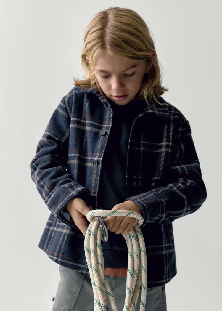 A young boy with blonde hair looks down while holding a coiled white rope with green and gray stripes. He is wearing a dark navy blue plaid flannel shirt over a black t-shirt and gray pants, against a plain light gray background.