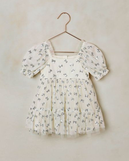 Beige dress with blue bow sequins on it on a hanger against a beige wall