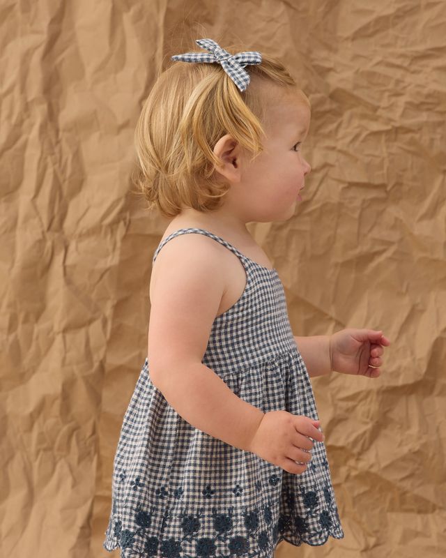 Young child wearing a checkered dress with a bow in their hair against a brown textured background