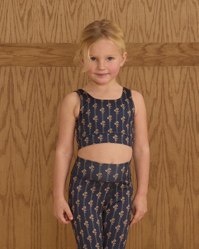 Young girl wearing a navy blue floral top and pants against a wooden paneled wall.