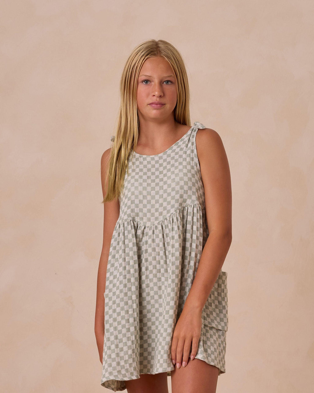 girl modeling the sage and beige check dress, sleeveless with two large pockets on either side of the dress. dress hits above the knee
