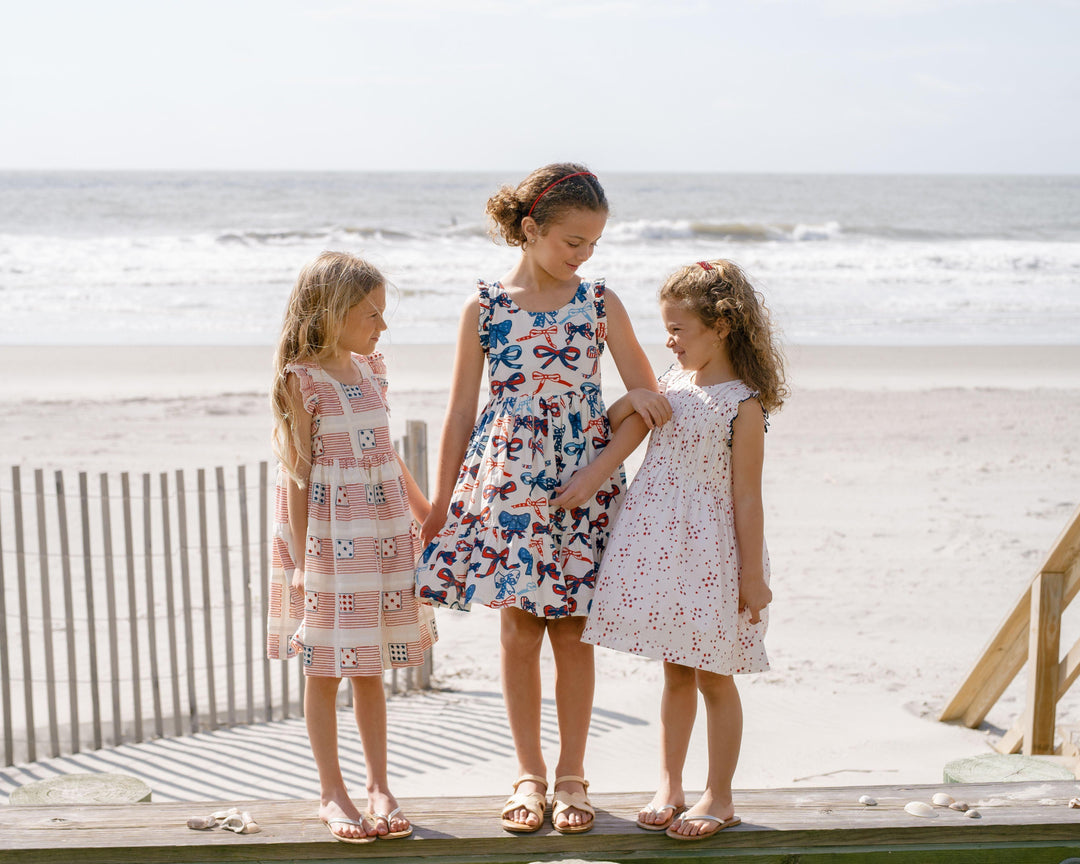 3 young girls dressed in Summer dresses. the girl in the middle dress has bows that have stars and stripes in the bow. 