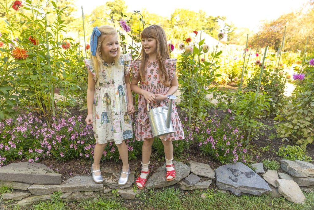 Two young girls standing in a garden with flowers and plants around them.