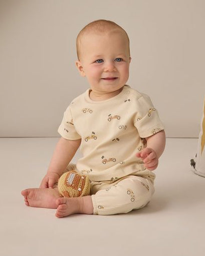 Baby sitting on a beige floor wearing a cream-colored outfit with small patterns, surrounded by wooden toys.