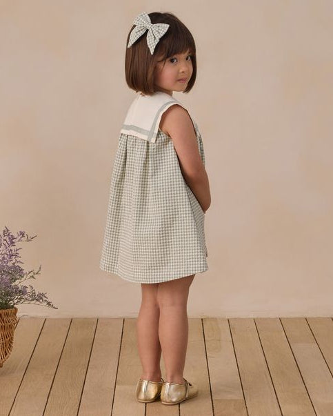 Young girl in a checkered dress standing on a wooden floor with a beige wall background