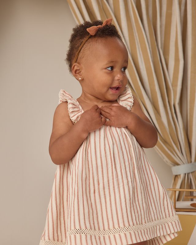 Young girl wearing a pink and white striped dress with ruffled sleeves, standing in front of a beige curtain.